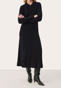 Robe en tricot noire à manches longues avec col et fermeture éclair, présentant une silhouette droite et arrivant mi-mollet. Portée avec des bottes hautes jusqu'aux genoux.