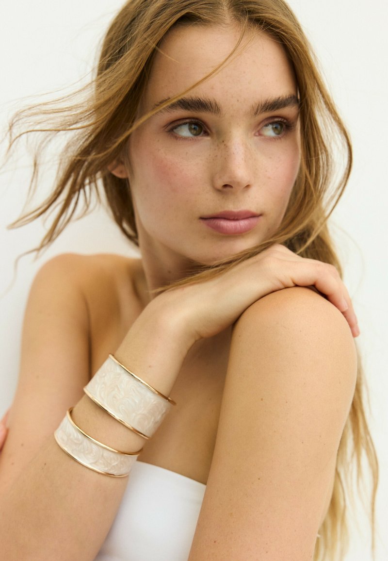 Jeune femme aux longs cheveux regardant sur le côté, reposant la main sur l'épaule, portant deux larges bracelets blancs et dorés sur le bras.