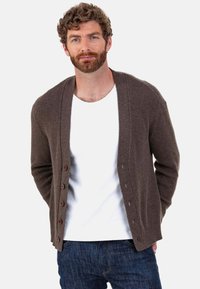 Cardigan marron c ôtelé avec un col châle, doté de boutons en bois sur le devant, porté par-dessus une chemise blanche et un jean bleu foncé.