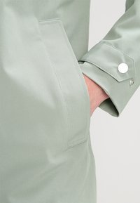 Hand teilweise in der Tasche eines hellgrünen Langarmshirts mit knöpfbarem Manschettendetail vor neutralem Hintergrund.