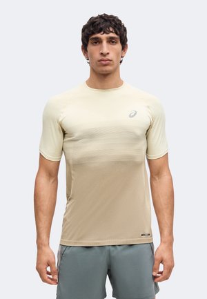 Man draagt een beige sportshirt met korte mouwen en grijze korte broek, staand tegen een effen lichte achtergrond, kijkend naar voren.