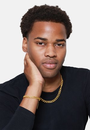 Gouden ketting en armband gedragen door een persoon met een gemiddelde huidskleur en kort, krullend haar, tegen een witte achtergrond. Zwart langarmshirt.