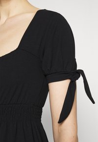 Vestido preto com decote quadrado, mangas bufantes com detalhes em laço e uma cintura ajustada com fru-fru para um formato ajustado. Tecido macio e elástico.