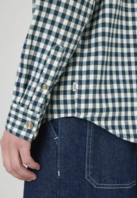 Chemise en flanelle à carreaux bleu marine et blanc avec des poignets boutonnés ; présente un ourlet droit et une petite étiquette logo près de la couture. Portée sur un jean denim foncé.