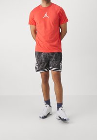Rotes T-Shirt mit weißem Logo, kombiniert mit schwarzen Shorts mit Blitzmuster und weißen Akzenten. Weiße Sneaker mit mehrfarbigen Details.