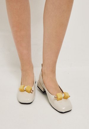 Chaussures blanches à petits talons portées sur des jambes nues avec de grosses décorations de perles géométriques jaunes à l'avant.