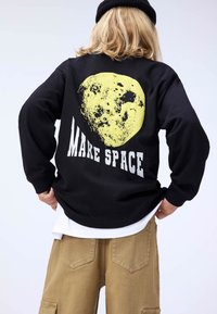 Black sweatshirt featuring a large yellow moon graphic and the text "MAKE SPACE" in bold white on the back. Layered over a white shirt.