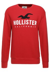 Sweatshirt en coton rouge avec une texture côtelée, des manches longues et un logo d'oiseau noir avec le texte "HOLLISTER CALIFORNIA" en blanc.