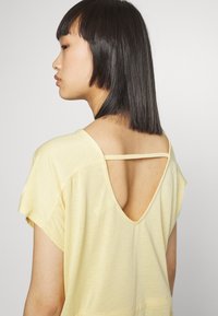 Top amarelo claro em tecido suave com costas em V. Apresenta mangas curtas e um detalhe de alça fina nas costas superiores.