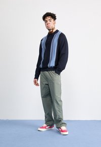 Jeune homme atteint de vitiligo portant un pull à rayures noir et bleu, un pantalon vert et des baskets roses, debout devant un fond blanc uni.