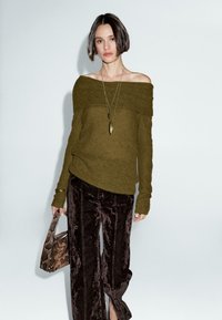 Olivgrüner, gestrickter Off-Shoulder-Pullover mit langen Ärmeln, kombiniert mit braunen, samtigen weit geschnittenen Hosen und einer geprägten Handtasche. Langer Halsketten-Akzent.
