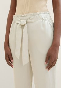 Ljust beige högmidjade byxor med ett tygbälte knutet fram. Slät textur med en lös passform och veckad midja.