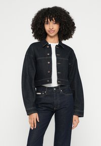 Krótka kurtka dżinsowa w ciemnym kolorze niebieskim z dużymi guzikami, szerokimi rękawami i klasycznym kołnierzem. Noszona na białej koszuli i zestawiona z ciemnymi dżinsami.