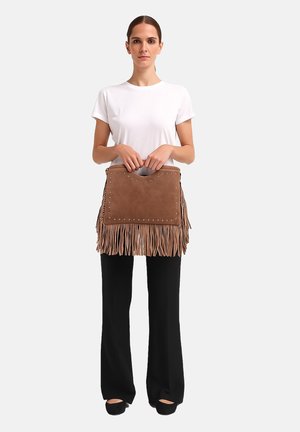 Vrouw in een wit T-shirt en zwarte broek die een bruin suède franjeshandtas met metalen studs vasthoudt voor een effen achtergrond.