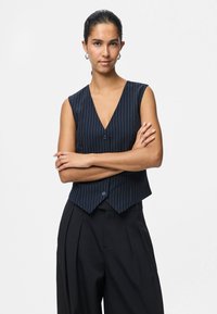 Sieviete tumšiem matiem sasietiem atpakaļ, ģērbusies bezpiedurkņu tumši zila svītrainā veste un brīvās melnās bikses, stāv ar rokām saliktām uz krūtīm pret balto fona.