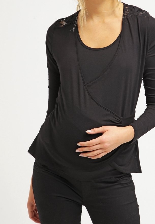 Femme enceinte portant un haut noir à manches longues avec des détails en dentelle sur les épaules, tenant son ventre d'une main.