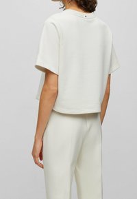 Kobieta ubrana w biały krótki top z krótkim rękawem i pasujące białe spodnie z wysokim stanem, widoczna od tyłu na jednolitym tle.