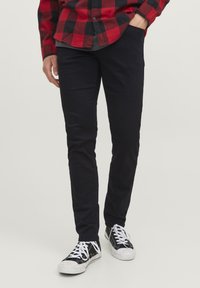 Pantalons slim-fit noirs avec une texture lisse, associ�és à des baskets noir et blanc. L'arrière-plan est neutre, mettant en valeur les détails des vêtements.