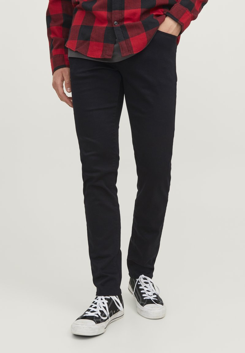 Pantalons slim-fit noirs avec une texture lisse, associés à des baskets noir et blanc. L'arrière-plan est neutre, mettant en valeur les détails des vêtements.