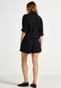 Set in lino nero composto da camicia e pantaloni corti con maniche arrotolate, vita aderente, tasche laterali e scarpe nere a tacco piatto, visto da dietro.