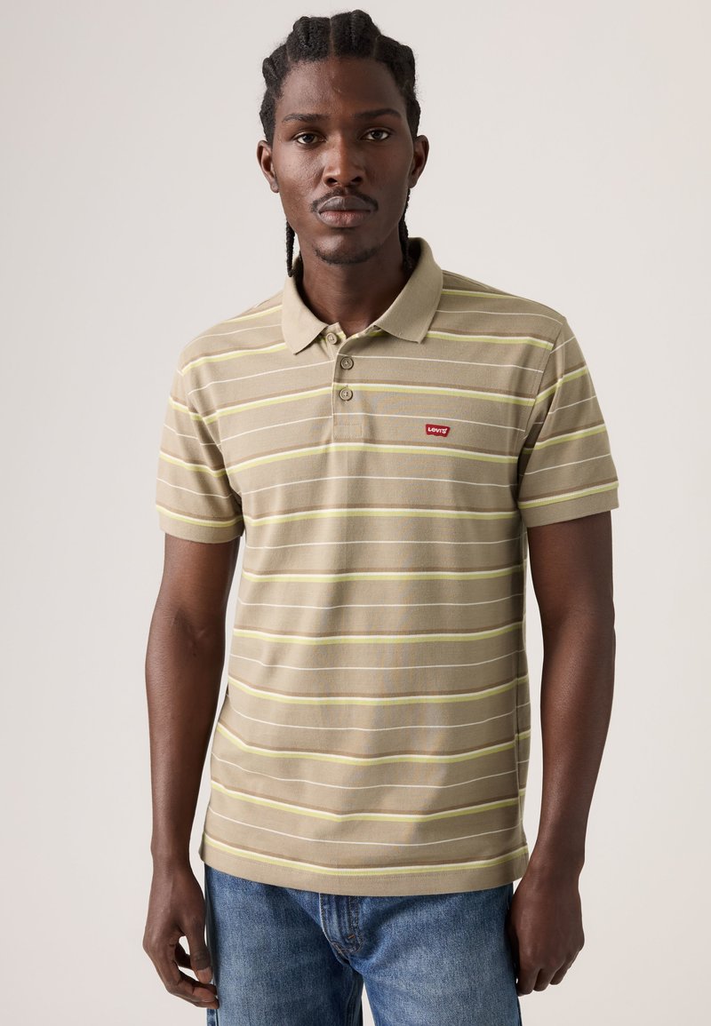 Man with braided hair wearing a beige and yellow striped Levi's polo shirt and blue jeans, standing against a plain background.