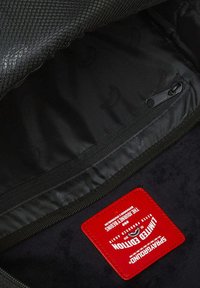 Interior de mochila negra con textura suave, panel de malla, bolsillo lateral con cremallera y una etiqueta roja con logo y texto.
