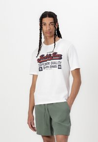 Young man with long braided hair wearing a white "Jack & Jones" graphic t-shirt and green shorts standing against a plain white background.