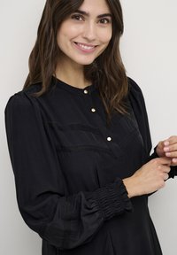 Zwarte blouse met lange mouwen, featuring een knopenlijst, textuurlijnen en geplooide manchetten. Glad materiaal met een subtiele glans.