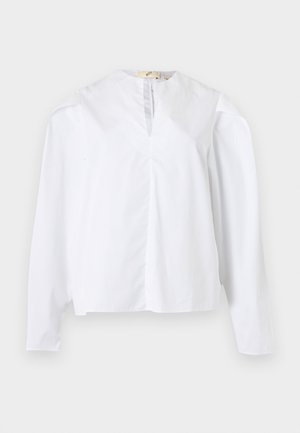 Woman with short hair wears white long-sleeve top with shoulder details and beige wide-leg pleated trousers, standing on white background.