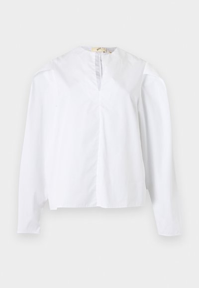 Blouse blanche à manches longues avec encolure ronde, fente à l'avant et coupe ample, présentée sur un fond clair uni.