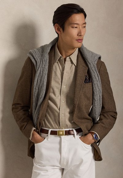 Moški v rjavi suknjič, bež srajci, belih hlačah, sivem puloverju prek ramen, rjavem pasu in ročni uri, zre stran.