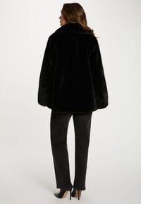 Cappotto in pelliccia sintetica nera con un ampio colletto, vestibilità oversize e maniche lunghe; indossato con jeans neri a gamba dritta e scarpe col tacco nere.