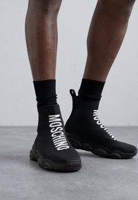Black sock-style sneakers with a chunky sole. Features white Moschino text on the side, seamless upper, and a ribbed ankle cuff.