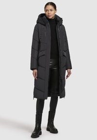 Dlouhý černý péřový kabát s kapucí, s prošívaným designem, zipovými kapsami a rovný střih, v kombinaci s černými skinny kalhotami a botami.