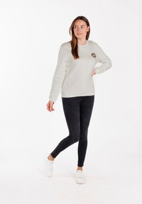 Felpa grigia chiara con scollo rotondo, polsini e orlo a coste. Presenta una toppa con stampa leopardata sul lato sinistro del petto. Indossata con jeans skinny neri e sneakers bianche.