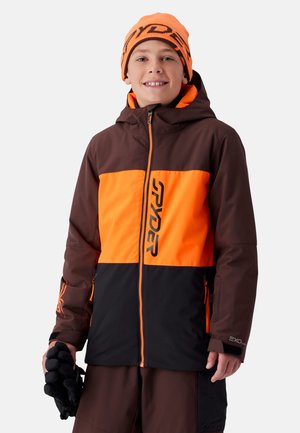 Garçon souriant portant une veste d'hiver marron et orange, un bonnet orange assorti et tenant des gants noirs dans une main.