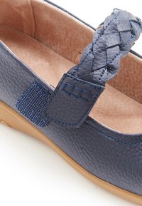 Zapato plano de cuero azul marino con un diseño de tira trenzada, interior acolchado y superficie texturizada. Presenta una suela de goma en contraste color marrón claro.