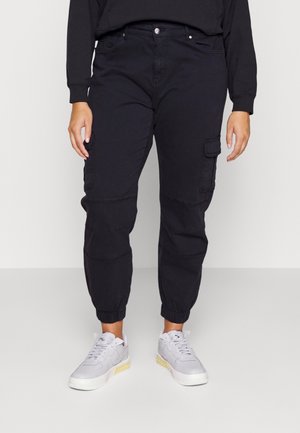 Person wearing black cargo pants with elastic cuffs and white sneakers, standing against a plain light background.