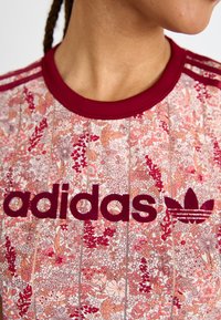 Ziedu raksta sporta krekls ar bordo akcentiem. Iespējo ribota apkakle, "adidas" logo un ikoniskās trīs svītras uz pleciem.