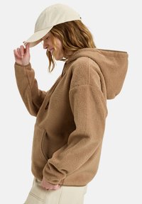 Brun fleece hoodie med en stor hætte, snoretræk og kengurulomme; parret med en beige kasket. Blød tekstur og afslappet pasform.