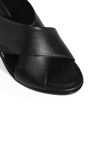Zwarte leren sandaal met gekruiste banden, open teen en een gladde textuur. Kenmerkt zich door een gesloten hiel en een platte zool.