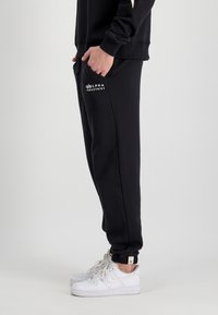 Čierne tepláky vyrobené z mäkkej látky, s elastickým pásom a manžetami, a bielym logom "Alpha Industries" na vrecku.