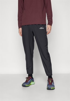Man draagt zwarte Nike trainingsbroek, bordeauxrode longsleeve en meerkleurige hardloopschoenen met groene zolen, staand tegen een effen achtergrond.