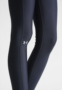 Leggings atléticos negros hechos de una tela suave y elástica. Las características incluyen costuras en contraste y un pequeño logo blanco en la parte inferior de la pierna.