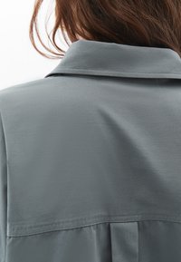 Blusa grigia con una texture liscia, caratterizzata da un colletto morbido a pieghe e un dettaglio plissettato nella parte posteriore. Il tessuto ha una leggera lucentezza.