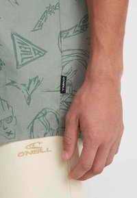 Groen met patroon shirt met lichtblauwe ontwerpen; de stof heeft een label met "O'Neill." Beige shorts met geborduurd "O'Neill" logo aan de zoom.