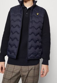 Marinblå vadderad väst med hög krage, med zip-öppning och en gul logotyp. Bärs över en strukturerad marinblå tröja och rutiga byxor.