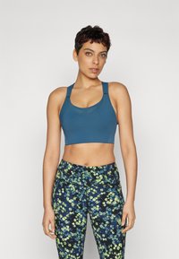 Tyrkis sports-bh med rund hals og brede stropper, kombinert med tettsittende leggings med mørk bakgrunn og lys blomstermønster.