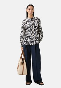 Langemouwen shirt met navy abstract patroon op crèmekleurige stof, gecombineerd met navy wijde broek en een grote beige geweven totebag met bruine accenten.