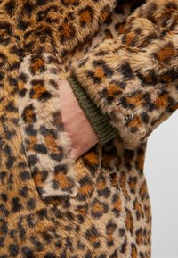 Leketmønsteret faux fur-kåpe med en myk tekstur, brune og svarte flekker, og grønne ribbede mansjetter med en sidelomme.